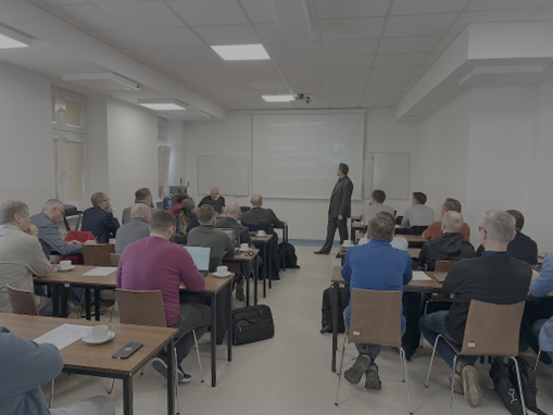 Grupa ludzi siedząca w sali. W tle mężczyzna omawiający coś na podstawie obrazu z projektora